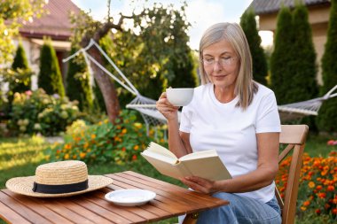 Bahçedeki ahşap masada içki içip kitap okuyan yaşlı bir kadın.