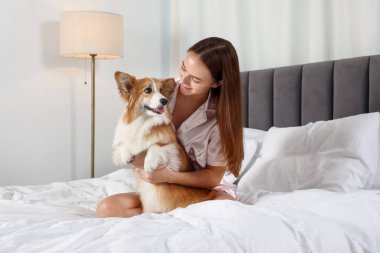 Evcil hayvan dostu otel. Güzel Galli Corgi köpeğiyle yatakta gülümseyen kadın.