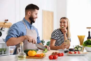 Yemek pişirme süreci. Karısı mutfaktaki masada akıllı telefondan yemek tarifine bakarken gülümseyen adam salatalık kesiyor.