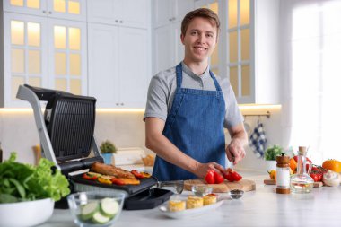 Adam mutfakta elektrikli ızgarada et ve sebze pişirirken biber kesiyor.