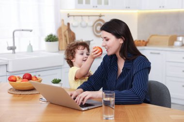 Gülümseyen anne laptopla çalışırken oğlu ev ofisinin ahşap masasında ona elma veriyor.