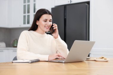 Dahili ofisteki ahşap masada bilgisayarla çalışırken akıllı telefondan konuşan gülümseyen kadın.