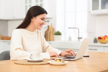 Ev ofisinin ahşap masasında dizüstü bilgisayarla çalışan gülümseyen kadın.