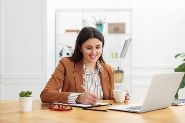 Takım elbiseli, güler yüzlü bir kadın ofis laptopuyla ahşap masada çalışıyor.