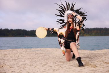 Kızılderili başlıklı güzel bir şaman kadın. Yüzü boyalı ve tef çalıyor. Sahilde nehir kenarında ayin yapıyor. Metin için boşluk