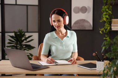 Kulaklıklı bir kadın, bilgisayarla kapalı bir masada çevrimiçi sınavdan geçerken not alıyor.