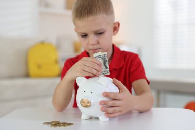 Küçük çocuk, içerideki beyaz masadaki kumbaraya cep harçlığı koyuyor.