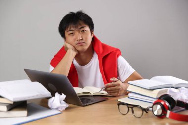 Dizüstü bilgisayarı ve kitapları olan öğrenci gri duvarın yanındaki tahta masada sınava hazırlanıyor.