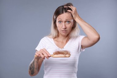 Alopecia sorunu. Gri arka planda saçları dökülmüş, elinde fırça tutan endişeli kadın.