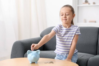 Küçük kız cep harçlığını ahşap masadaki domuz kumbarasına yatırıyor.