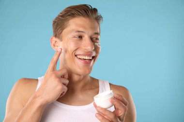 Yakışıklı genç adam açık mavi arka planda yüzüne krem sürüyor.