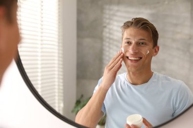 Yakışıklı genç adam evde aynanın yanında yüzüne krem sürüyor.