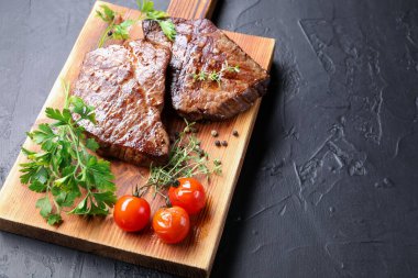 Delicious grilled beef steaks with herbs, peppercorns and tomatoes on dark textured table, closeup. Space for text