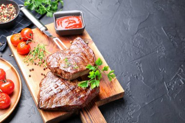 Delicious grilled beef steaks with herbs, peppercorns, tomatoes, carving fork and ketchup on dark textured table, closeup. Space for text