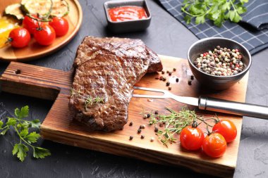 Delicious grilled beef steak with herbs, peppercorns, tomatoes, carving fork and ketchup on dark textured table, closeup
