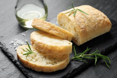 Cut crispy ciabatta, rosemary, salt and oil on black textured table, closeup