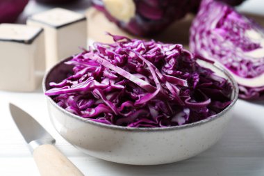 Shredded red cabbage in bowl and knife on white wooden table, closeup