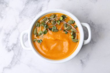 Delicious pumpkin soup with seeds and microgreens in bowl on white marble table, top view
