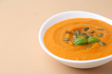 Delicious pumpkin soup with seeds and basil in bowl on beige table, closeup. Space for text