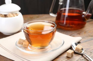 Aromatic black tea in glass cup, brown sugar and teapot on wooden table, closeup