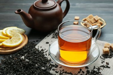 Aromatic black tea in glass cup, dried leaves, brown sugar, lemon and teapot on wooden table, closeup