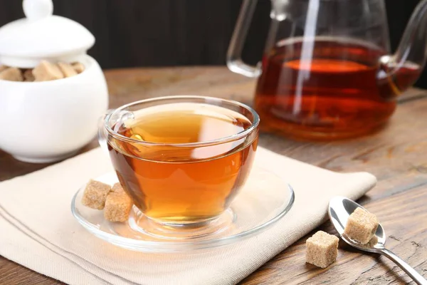 Aromatic black tea in glass cup, brown sugar and teapot on wooden table, closeup