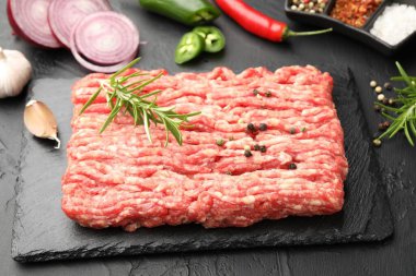 Raw minced meat with rosemary, spices, onion and chili peppers on black textured table, closeup