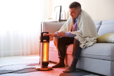 Evinde elektrikli ısıtıcının yanında battaniyeli genç bir adam var. Aygıttan çıkan turuncu sıcaklık ışığı