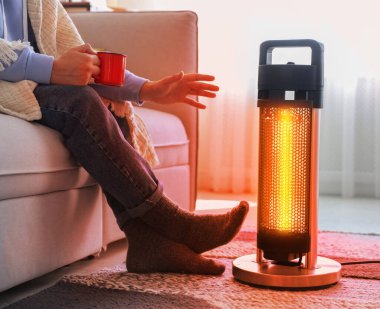 Evinde elektrikli ısıtıcının yanında fincanı olan genç adam, yakın çekim. Aygıttan çıkan turuncu sıcaklık ışığı