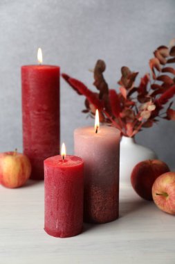Burning candles, apples and decorative branches in vase on light wooden table against gray background, closeup. Autumn atmosphere