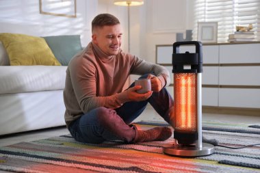 Evinde elektrikli ısıtıcının yanında fincanı olan genç bir adam. Aygıttan çıkan turuncu sıcaklık ışığı