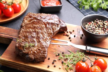 Delicious grilled beef steak with herbs, peppercorns, tomatoes, carving fork and ketchup on dark textured table, closeup