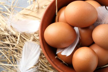 Raw chicken eggs in bowl, feathers and straw on table, top view