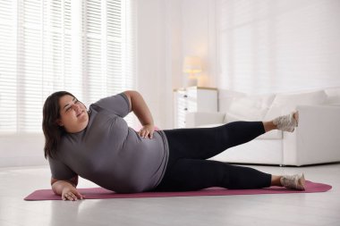 Evde yoga minderinde spor kıyafetleriyle gülümseyen şişman kadın.