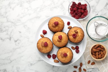 Fresh muffins with raspberries, almonds and flour on white marble table, flat lay. Space for text