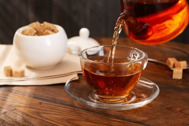 Pouring aromatic black tea into glass cup at wooden table, closeup