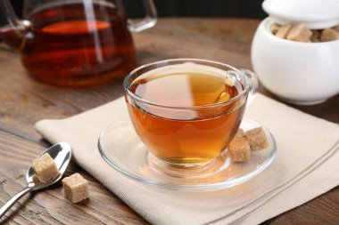 Aromatic black tea in glass cup, brown sugar and teapot on wooden table, closeup