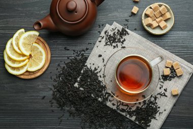 Aromatic black tea in glass cup, dried leaves, brown sugar, lemon and teapot on wooden table, flat lay