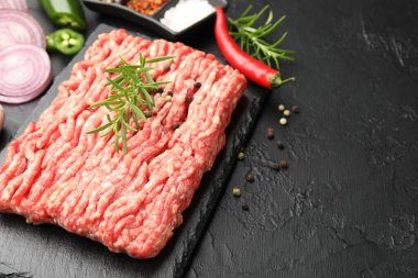 Raw minced meat with rosemary, spices, onion and chili peppers on black textured table, closeup. Space for text