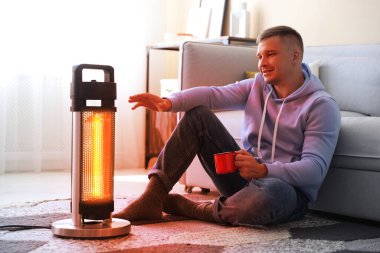 Evinde elektrikli ısıtıcının yanında fincanı olan genç bir adam. Aygıttan çıkan turuncu sıcaklık ışığı