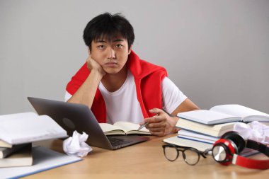 Dizüstü bilgisayarı ve kitapları olan öğrenci gri duvarın yanındaki tahta masada sınava hazırlanıyor.