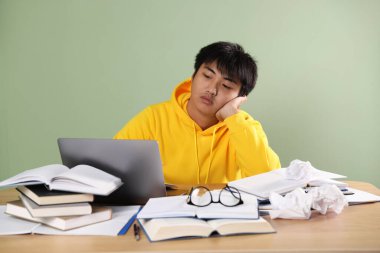 Laptoplu ve kitaplı öğrenci yeşil duvarın yanındaki tahta masada sınava hazırlanıyor.