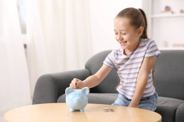 Küçük kız cep harçlığını ahşap masadaki domuz kumbarasına yatırıyor.