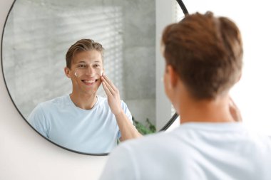 Yakışıklı genç adam evde aynanın yanında yüzüne krem sürüyor.