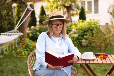 Bahçedeki ahşap masada kitap okuyan yaşlı bir kadın.