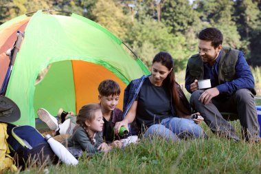 Aile, kamp çadırı ve bardaklar dolusu sıcak içecekle birlikte dışarıda vakit geçiriyor.