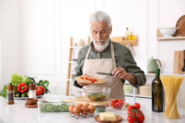 Yaşlı adam mutfaktaki beyaz mermer masada salata pişiriyor.