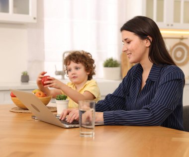 Oğlu ev ofisinin ahşap masasında elmayla oynarken dizüstü bilgisayarla çalışan gülümseyen kadın.