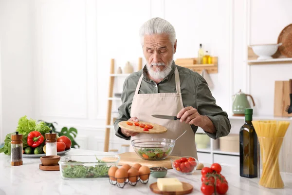 Yaşlı adam mutfaktaki beyaz mermer masada salata pişiriyor.
