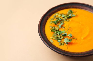 Delicious pumpkin soup with seeds and microgreens in bowl on beige table, closeup. Space for text